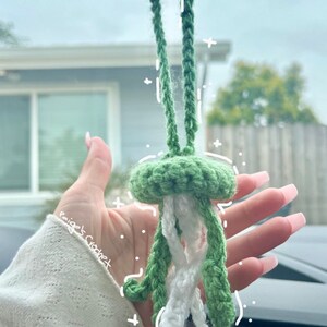 May include: A green crocheted jellyfish car charm with white tentacles hanging from the rearview mirror of a car. The charm is held in a hand with pink nail polish. The text "Paige Crochet" is written on the image.