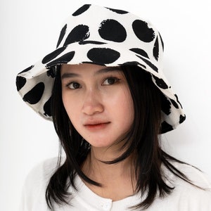 May include: A white bucket hat with large black polka dots. The hat is worn by a person with long black hair.