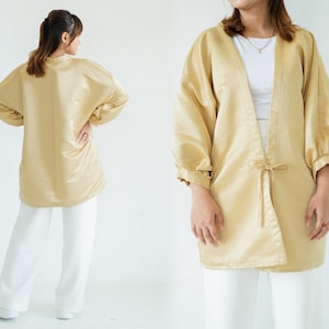 Puede incluir: Una chaqueta estilo kimono de satén amarillo dorado con mangas largas y voluminosas y un cierre de lazo en la cintura. La chaqueta se combina con pantalones blancos. La tela tiene un brillo sutil y el estilo general es relajado y elegante.