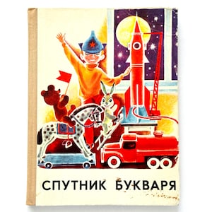 Op de afbeelding: Een vintage hardcover boek getiteld "СПУТНИК БУКВАРЯ" met een kleurrijke illustratie van een kind met een blauwe sterrenmuts, een bruine teddybeer op een gevlekt hobbelpaard, een konijn op een ladder, een rode raket en een rode tankwagen.