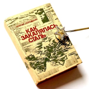 May include: A vintage paperback book titled "Как закалялась сталь" by Nikolai Ostrovsky. The book cover features a map and a red star bookmark.