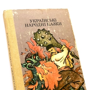 Pode incluir: Uma capa de livro vintage com uma ilustração colorida de um homem lutando contra um monstro com um porrete com pontas. O título do livro é "Українські народні казки", que se traduz como "Contos populares ucranianos".