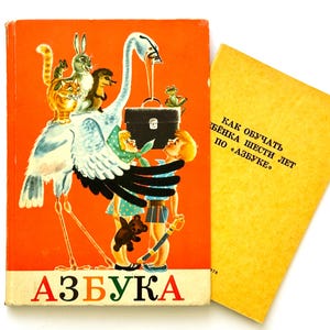 May include: A vintage orange book titled AZBUKA featuring a large white stork carrying a black briefcase with various animals and two children, alongside a yellow companion book with Russian text that reads KAK OBUCHAT REBENKA SHESTI LET PO AZBUKE.