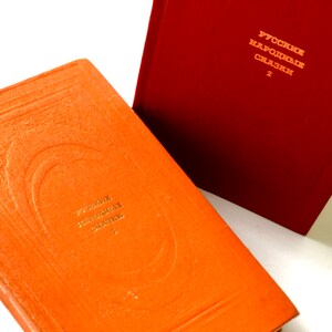 Pode incluir: Dois livros encadernados em laranja com letras douradas na lombada. O texto na lombada do livro superior diz "РУССКИЕ НАРОДНЫЕ СКАЗКИ 2". O texto na lombada do livro inferior diz "РУССКИЕ НАРОДНЫЕ СКАЗКИ 1".