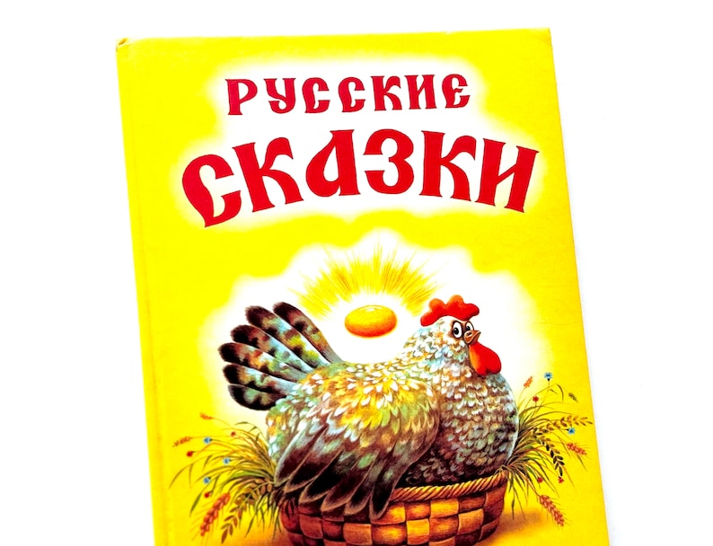 May include: A yellow book cover with the title "Русские сказки" in red letters. The cover features an illustration of a hen sitting in a basket with a golden egg.