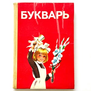 Pode incluir: Um livro de capa dura vermelho vintage intitulado BUKVAR com uma lombada de cor clara. A capa apresenta a ilustração de uma criança de cabelos loiros vestindo um avental branco sobre um vestido escuro, segurando um sino prateado e flores brancas.