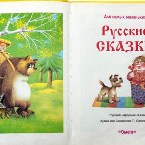 May include: A vintage children's book cover featuring a brown bear carrying a basket with a girl inside. The bear is standing in a forest setting with a green background. The book title is "Russian Fairy Tales" in large, bold letters. The book is for young children.