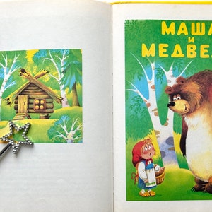 May include: A children's book cover with a colorful illustration of a girl in a red hood and a bear. The title of the book is "Masha and the Bear" in Russian.