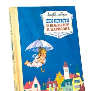 Può includere: Una copertina di libro blu con un titolo in russo che recita "Tre storie sul bambino e Karlsson." La copertina presenta un'illustrazione di un bambino con una camicia rossa e capelli gialli che tiene un ombrello, volando in aria sopra una città.