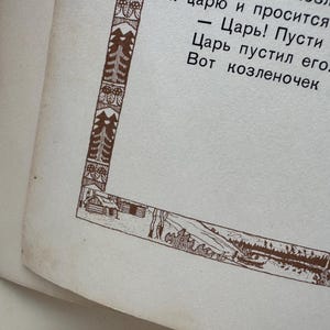 May include: Close-up of a vintage book page with text in a foreign language. The page has a decorative brown border featuring trees, a cabin, and a landscape scene. The paper is aged with a slightly yellowed tone.