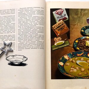 May include: A vintage cookbook open to a page with a recipe for soup. The page features illustrations of bowls of soup, a plate of buckwheat porridge, and a glass of juice. The cookbook also features advertisements for canned goods, including 'Kučero' and 'Гречневая'.