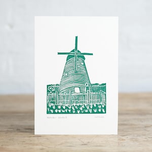 Könnte beinhalten: Ein weißer rechteckiger Druck mit einer petrolfarbenen Linolschnitt-Illustration einer Windmühle hinter einem Holzzaun und einem Blumenfeld. Der Text "Holme-next-Sea Windmill" ist unten links gedruckt, mit den Initialen "cw 2022" auf der rechten Seite.