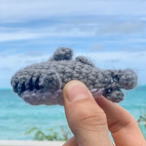 May include: A gray crocheted shark toy with a pink underbelly. The toy is being held in a hand against a blue sky and ocean background.