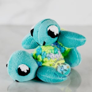 May include: Two crocheted plush sea turtles, one turquoise blue and one green, with black eyes and white accents. The turtles are stacked on top of each other. The larger turtle has a crocheted shell with green, yellow, and purple stripes. The smaller turtle is lying on its back with its legs in the air. The tag on the larger turtle reads "SS Handcraft Home of the Marble Turtle".