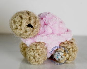 May include: A small crocheted turtle with a pink fuzzy shell and brown crocheted legs and head. The turtle is sitting on a white surface.