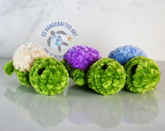 May include: Four crocheted turtle toys in different colors: cream, purple, blue, and green. The turtles are lying on a white surface. The text "SS HANDCRAFTED ART HOME OF THE MARBLE TURTLE" is visible in the background.