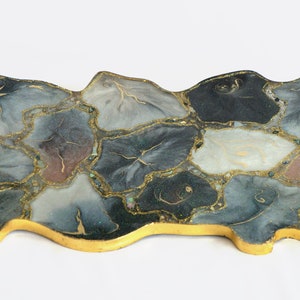 Peut inclure: Plateau de service décoratif avec un design unique. Le plateau présente un bord et des poignées dorés, et une surface en forme de mosaïque avec des nuances de gris, de bleu et de marron, accentuée de veines dorées. Un article de décoration élégant.