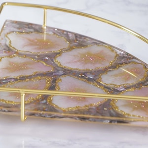 Peut inclure: Un plateau d'angle décoratif avec un cadre en métal doré. La surface du plateau présente un motif marbré avec des accents roses et dorés, ressemblant à des tranches d'agate. Le plateau est conçu pour être placé dans un coin.