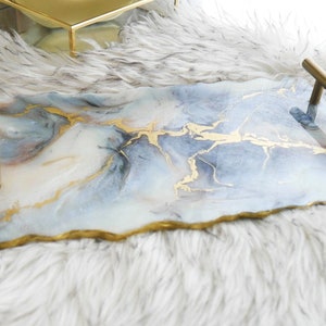 Puede incluir: Una bandeja rectangular con un diseño veteado en tonos azules, blancos y dorados. La bandeja tiene asas doradas y un borde pintado de oro. La bandeja descansa sobre una superficie blanca y esponjosa.