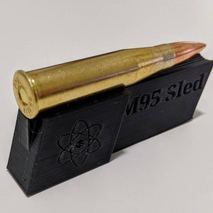 May include: A brass bullet rests on a black 3D-printed stand. The bullet has a copper-colored tip and a gold casing. The stand is angled and features the text "M95 Sled" and a stylized atom symbol.