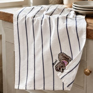 May include: A white kitchen towel featuring vertical navy blue stripes. The towel includes an embroidered graphic of a small brown dog wearing a pink collar, positioned next to the name Maxi stitched in black cursive lettering on the fabric.