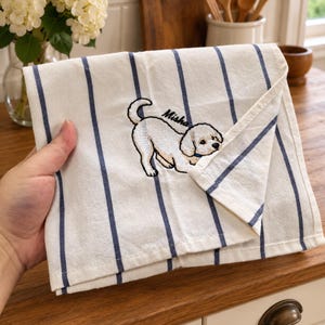 Pode incluir: Um pano de cozinha branco com riscas verticais azul-marinho e um bordado que mostra um cão pequeno de cor clara numa postura brincalhona. O nome Misha está bordado em letras cursivas pretas por cima da figura do cão no tecido.