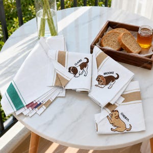 Pode incluir: Um conjunto de panos de prato de algodão branco com ilustrações de cães bordadas e os nomes Claire, Brooke e Cheryl. Os panos têm bordas listradas em tons de bege e verde, dispostos ao lado de uma bandeja de madeira com pão fatiado.