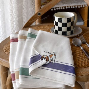 Pode incluir: Uma pilha de quatro panos de prato brancos com listras horizontais em vermelho, bege, verde e roxo. O pano superior apresenta um cão bordado em castanho e branco com o nome Cooper bordado acima em fio preto.