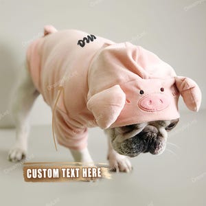 Könnte beinhalten: Ein hellrosa Haustier-Kapuzenpullover mit einem Schweinegesicht-Design auf der Kapuze, inklusive gestickten Augen, einer Schnauze und zwei abstehenden Ohren. Auf dem Rücken befindet sich eine schwarze Schreibschrift-Stickerei. Getragen von einem kleinen Hund.