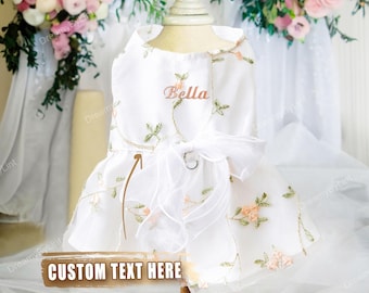 May include: A white dog dress with floral embroidery and a large bow. The dress features the name "Bella" embroidered on the bodice and the text "CUSTOM TEXT HERE" on a banner. The dress is displayed on a mannequin.