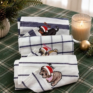 Pode incluir: Três panos de cozinha brancos dobrados com riscas azul-escuras e padrões xadrez. Cada pano apresenta um cão bordado com um gorro de Pai Natal vermelho, com os nomes Mochi, Snoosh e Mazi bordados sobre as ilustrações dos cães.