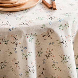 May include: A round, beige tablecloth with a delicate floral pattern in shades of green, orange, and blue. The tablecloth is made of a soft, linen-like fabric.