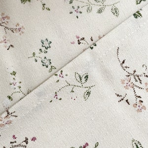 Pode incluir: Close-up de um tecido branco com um delicado padr&atilde;o floral em tons de verde, rosa e marrom. O tecido tem uma textura sutil e um fundo ligeiramente branco quebrado.