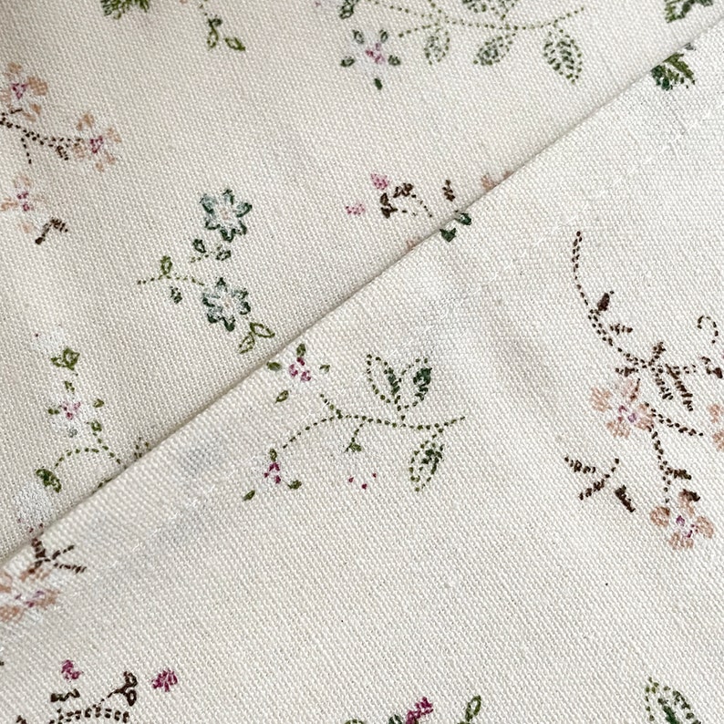Pode incluir: Close-up de um tecido branco com um delicado padr&atilde;o floral em tons de verde, rosa e marrom. O tecido tem uma textura sutil e um fundo ligeiramente branco quebrado.