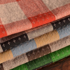 May include: A stack of folded fabric featuring a checkered pattern in shades of red, black, blue, yellow, brown, and green. Each square block of colour is accented with a small, white, cross-stitch embroidery detail throughout the textile surface.