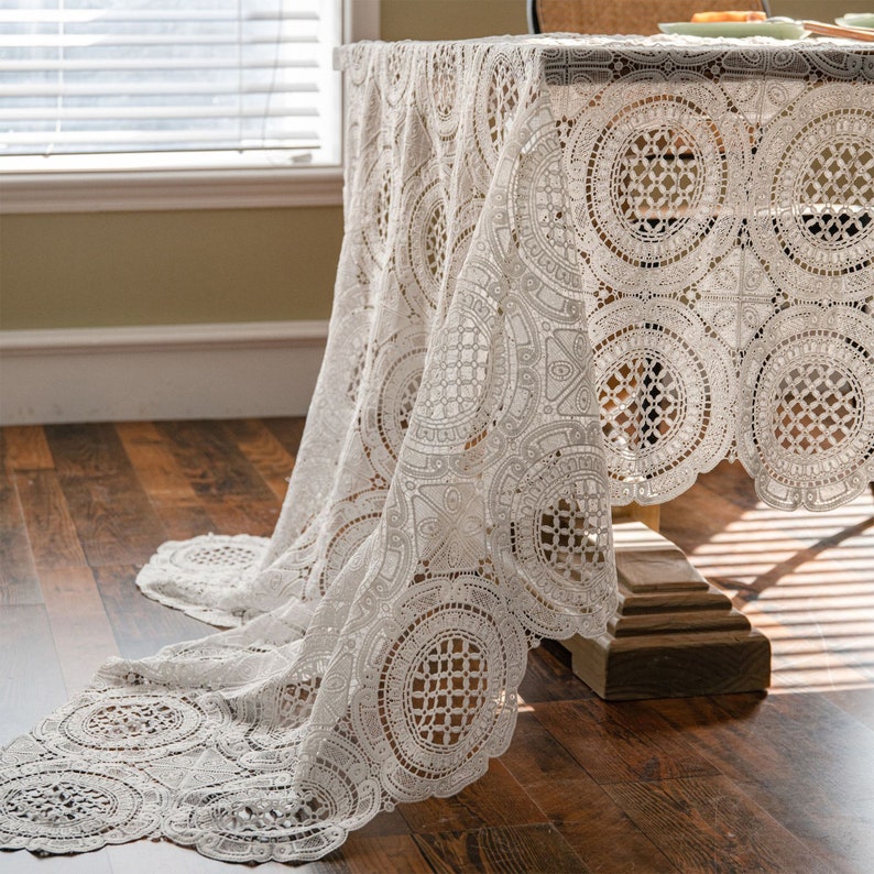 May include: White crochet tablecloth with a delicate circular pattern. The tablecloth is draped over a wooden table and hangs down to the floor.