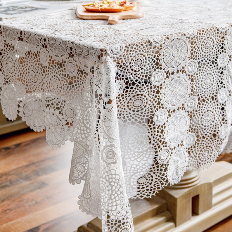 May include: White crochet tablecloth with a delicate floral pattern. The tablecloth is draped over a wooden table and has a scalloped edge.