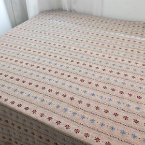 May include: A beige fabric tablecloth featuring a repeating pattern of horizontal stripes. The design consists of alternating rows of small, dark red six-petaled flowers and light blue six-petaled flowers, separated by thin, textured lines.