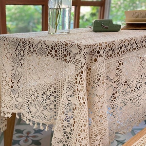 K&ouml;nnte beinhalten: Eine cremefarbene geh&auml;kelte Tischdecke mit einem zarten Blumenmuster und Fransenbesatz. Die Tischdecke ist &uuml;ber einen Holztisch mit Fliesenboden gelegt.