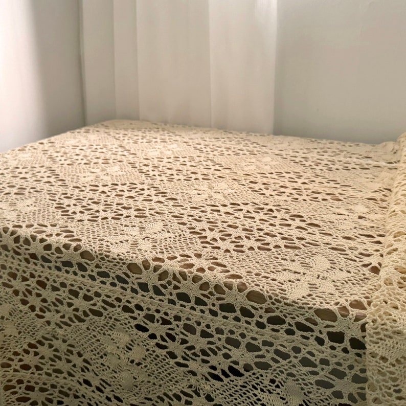 K&ouml;nnte beinhalten: Eine cremefarbene H&auml;keltischdecke mit einem zarten Blumenmuster. Die Tischdecke ist aus feinem Garn gefertigt und hat eine weiche, luftige Textur.