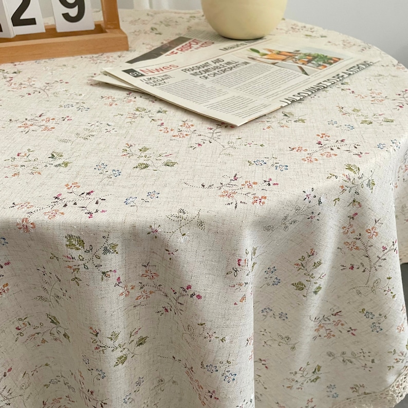 Pode incluir: Uma toalha de mesa redonda branca com um delicado padr&atilde;o floral em tons de rosa, azul e verde. A toalha de mesa tem uma borda de renda.