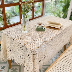 K&ouml;nnte beinhalten: Eine wei&szlig;e H&auml;keltischdecke mit einem zarten Blumenmuster und Fransenbesatz. Die Tischdecke liegt auf einem Holztisch mit einer Blumenvase und einem Buch.