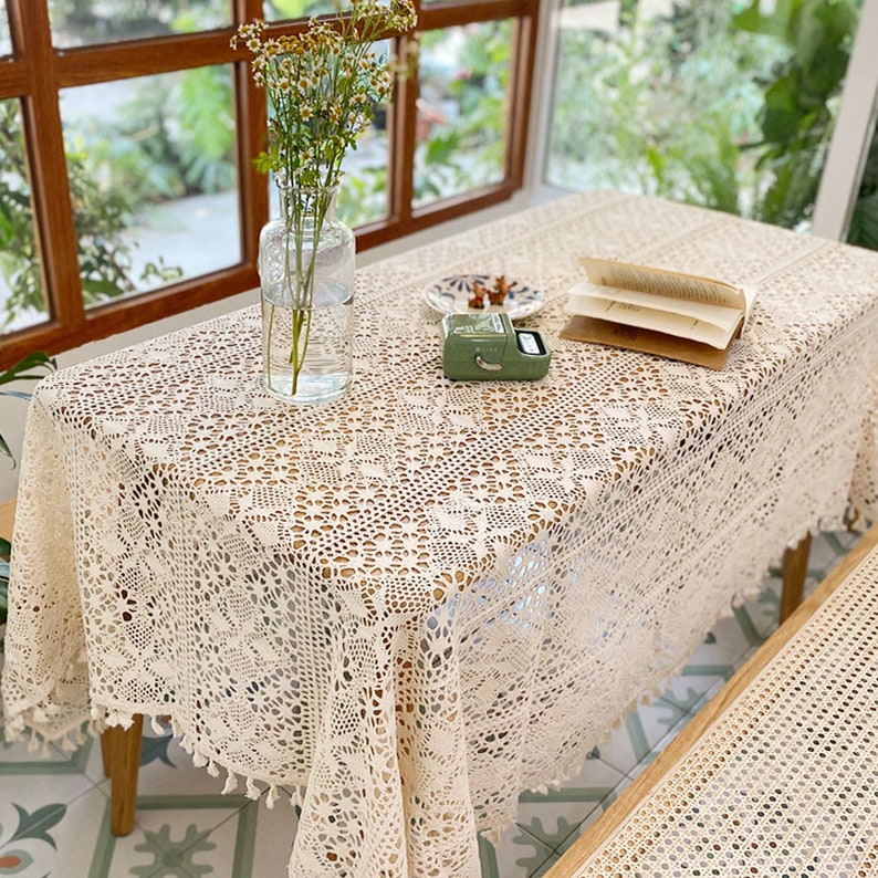 K&ouml;nnte beinhalten: Eine wei&szlig;e H&auml;keltischdecke mit einem zarten Blumenmuster und Fransenbesatz. Die Tischdecke liegt auf einem Holztisch mit einer Blumenvase und einem Buch.