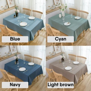 May include: Four tablecloths in different colors: blue, cyan, navy, and light brown. Each tablecloth is laid on a wooden table with two chairs. The tablecloths are all rectangular and have a simple design.