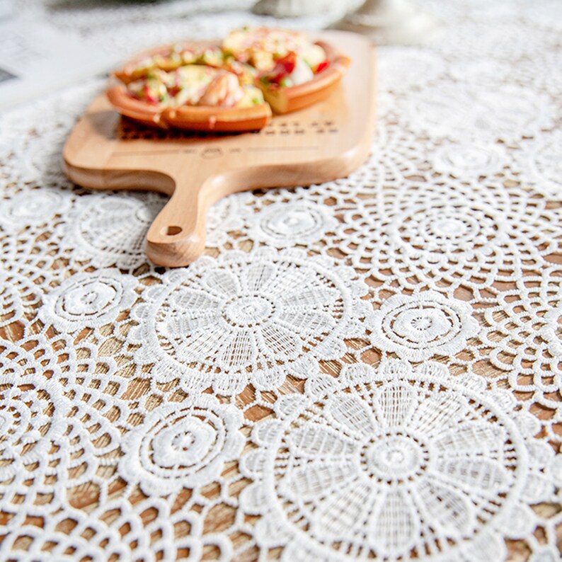 May include: White crocheted table runner with a delicate floral pattern. The runner is laid on a wooden table with a wooden cutting board and pizza slices on top.