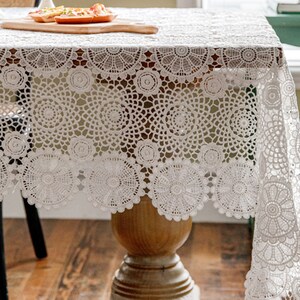 May include: White crochet tablecloth with a delicate floral pattern. The tablecloth has a scalloped edge and is draped over a wooden table.