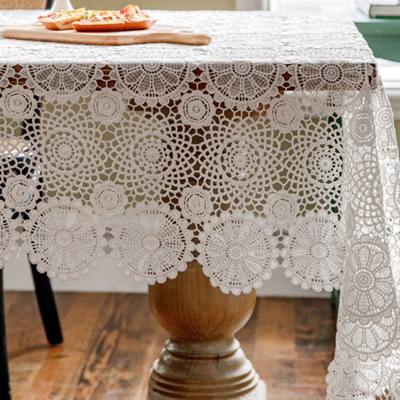May include: White crochet tablecloth with a delicate floral pattern. The tablecloth has a scalloped edge and is draped over a wooden table.