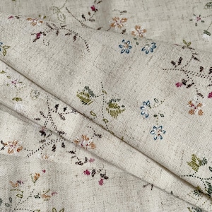 Pode incluir: Um close-up de um tecido bege com um delicado padr&atilde;o floral. O tecido apresenta pequenas flores bordadas em tons de rosa, azul, verde e marrom. O tecido tem uma textura sutil e uma sensa&ccedil;&atilde;o ligeiramente &aacute;spera.