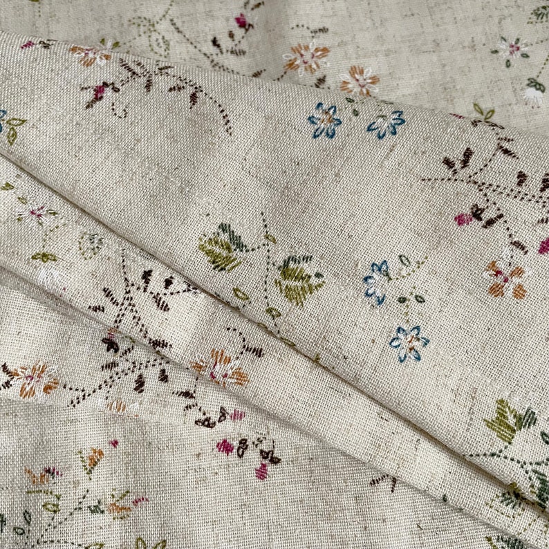 Pode incluir: Um close-up de um tecido bege com um delicado padr&atilde;o floral. O tecido apresenta pequenas flores bordadas em tons de rosa, azul, verde e marrom. O tecido tem uma textura sutil e uma sensa&ccedil;&atilde;o ligeiramente &aacute;spera.