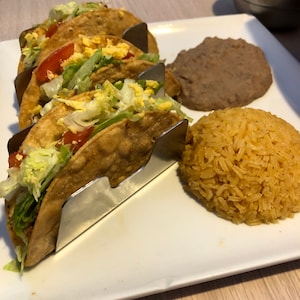 Pode incluir: Um prato branco com três tacos num suporte de tacos de metal, um acompanhamento de feijão refogado e um acompanhamento de arroz amarelo.